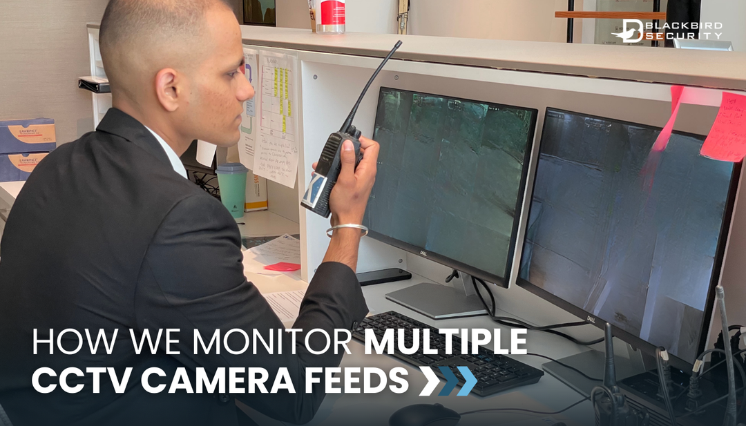 A security guard sits in front of a double monitor set up, monitoring CCTV footage. He's speaking into a walkie-talkie.