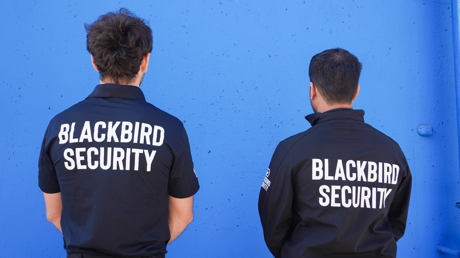 Two security officers facing a green wall outside a building in Regina. The back of their jackets read Blackbird Security.