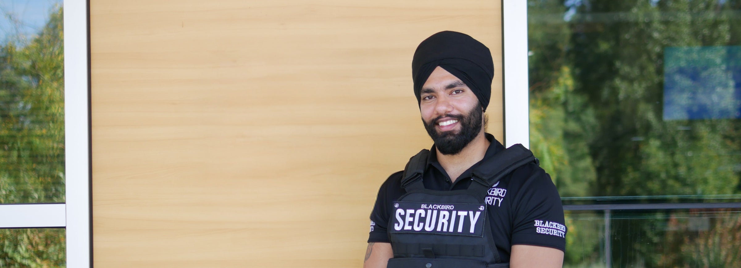 Man wearing a security shirt standing in front of a wooden door with trees in the background