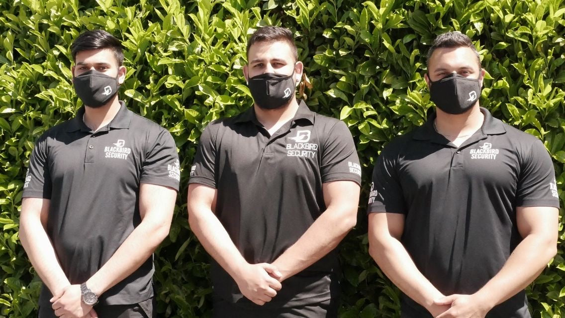 Three men wearing black polo shirts with logos and face masks standing in front of a green hedge.