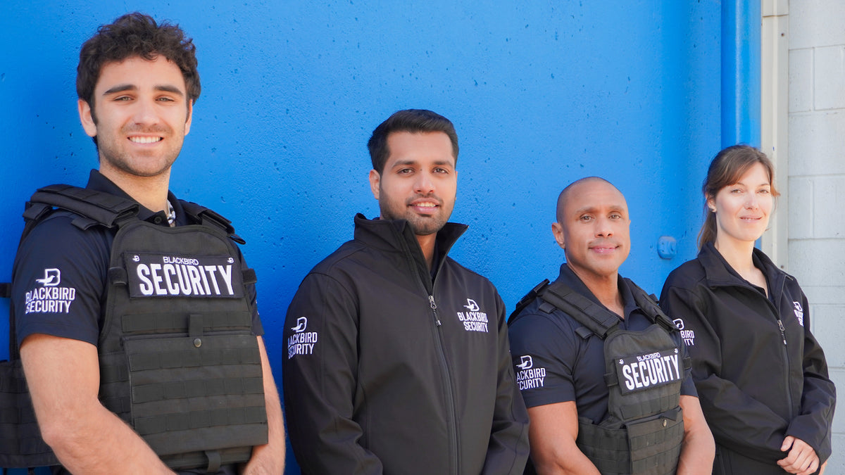 Four security guards stand together against a blue building. They are all in uniform, smiling with their hands gathered together at a relaxed pose