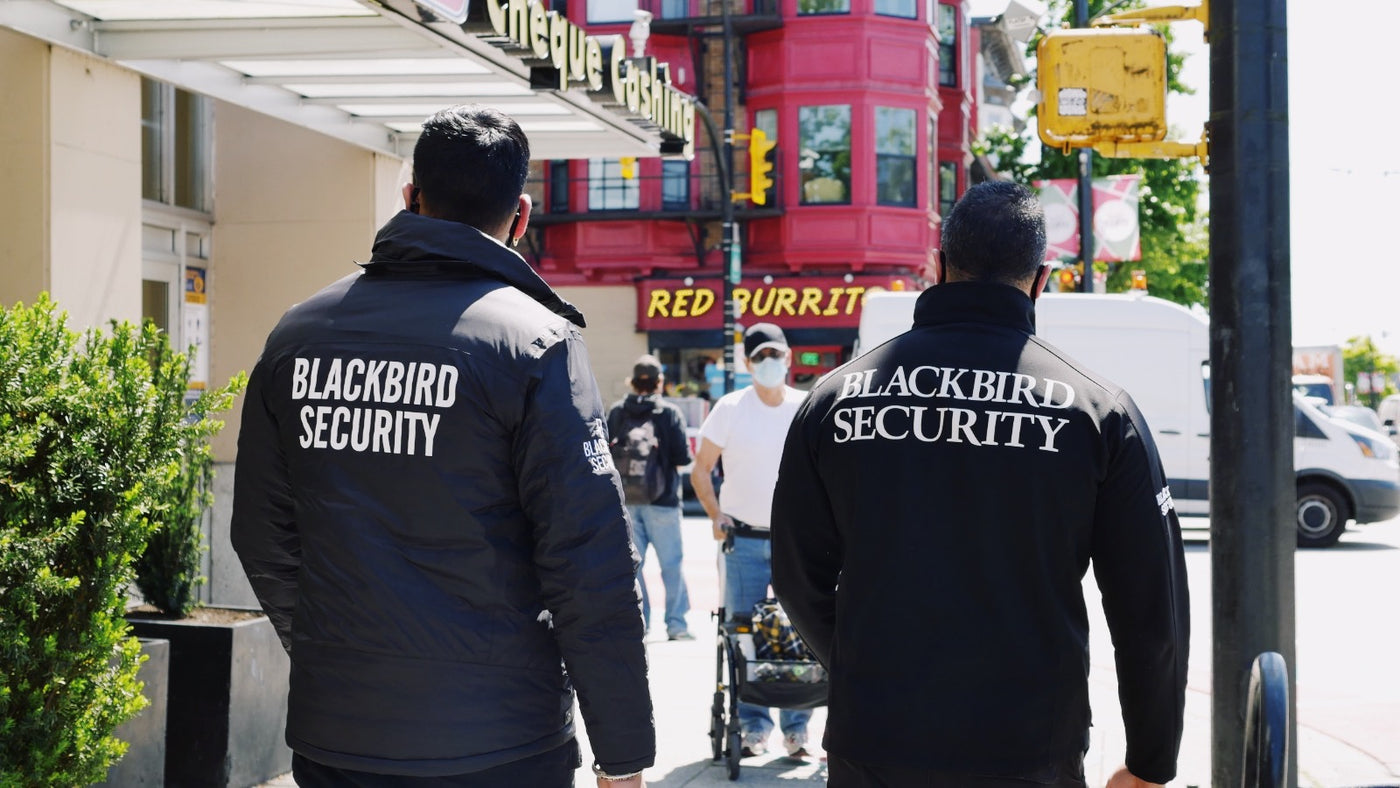 Two uniformed security guards walk down a busy street next two each other