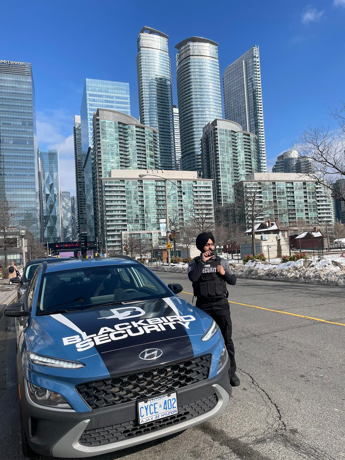 A tactical security guard stands outside of his mobile patrol security vehicle on the side of the road. A city backdrop full of skyscrapers is in the distance