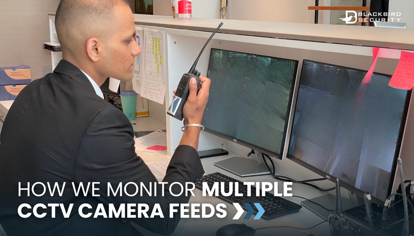 A security guard sits in front of a double monitor set up, monitoring CCTV footage. He's speaking into a walkie-talkie. 