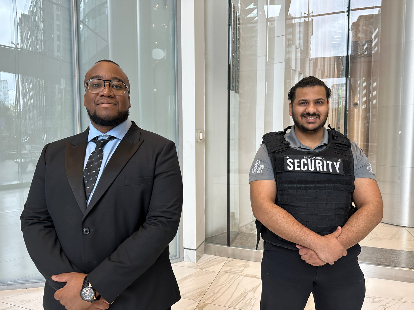 A uniformed security guard and a luxury security guard stand in front of a condominium building entrance smiling while providing residential security