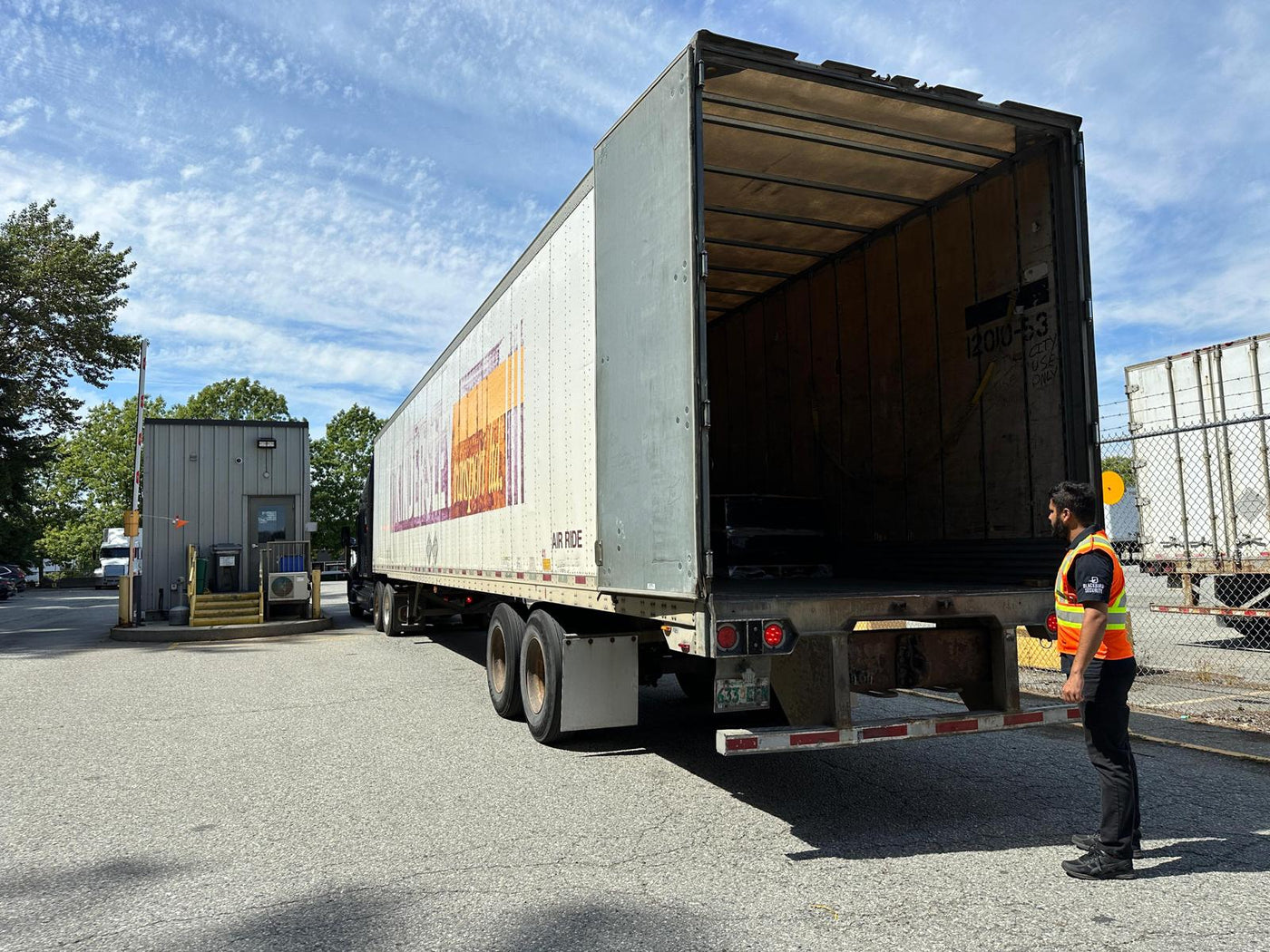 A uniformed security guard in a high-vis vest stands outside of a cargo truck that is open so that he can check the contents of the trailer