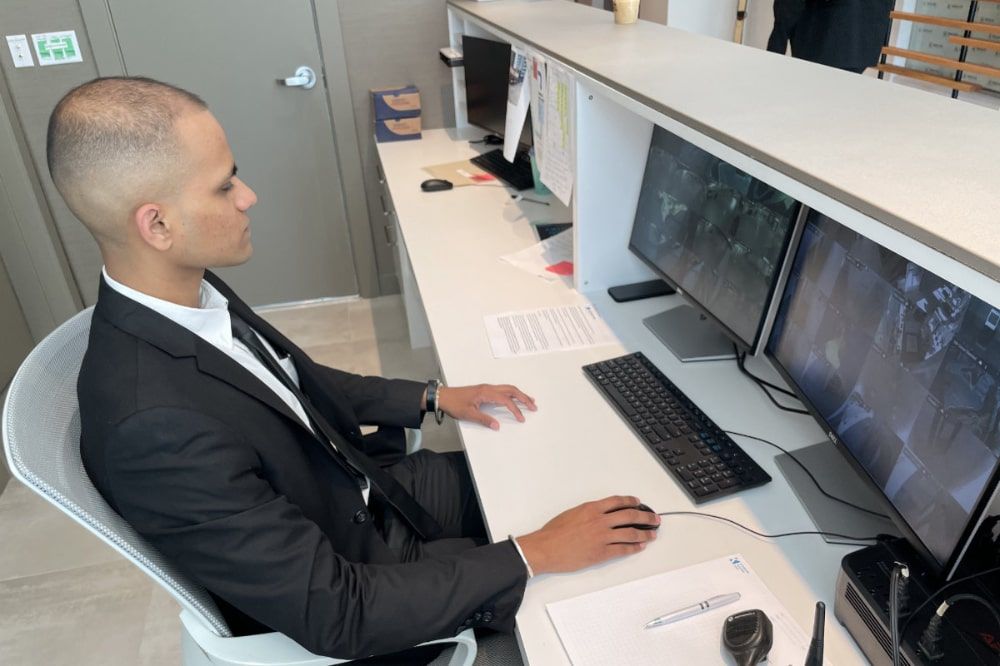 Uniformed Blackbird security guard sitting at concierge desk in front of two security camera screens