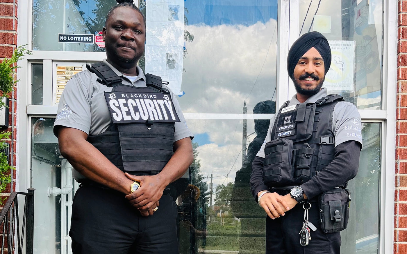 Two tactical security guards stand next to each other outside of a community building. Both of them are smiling and clasping their hands together in front of them