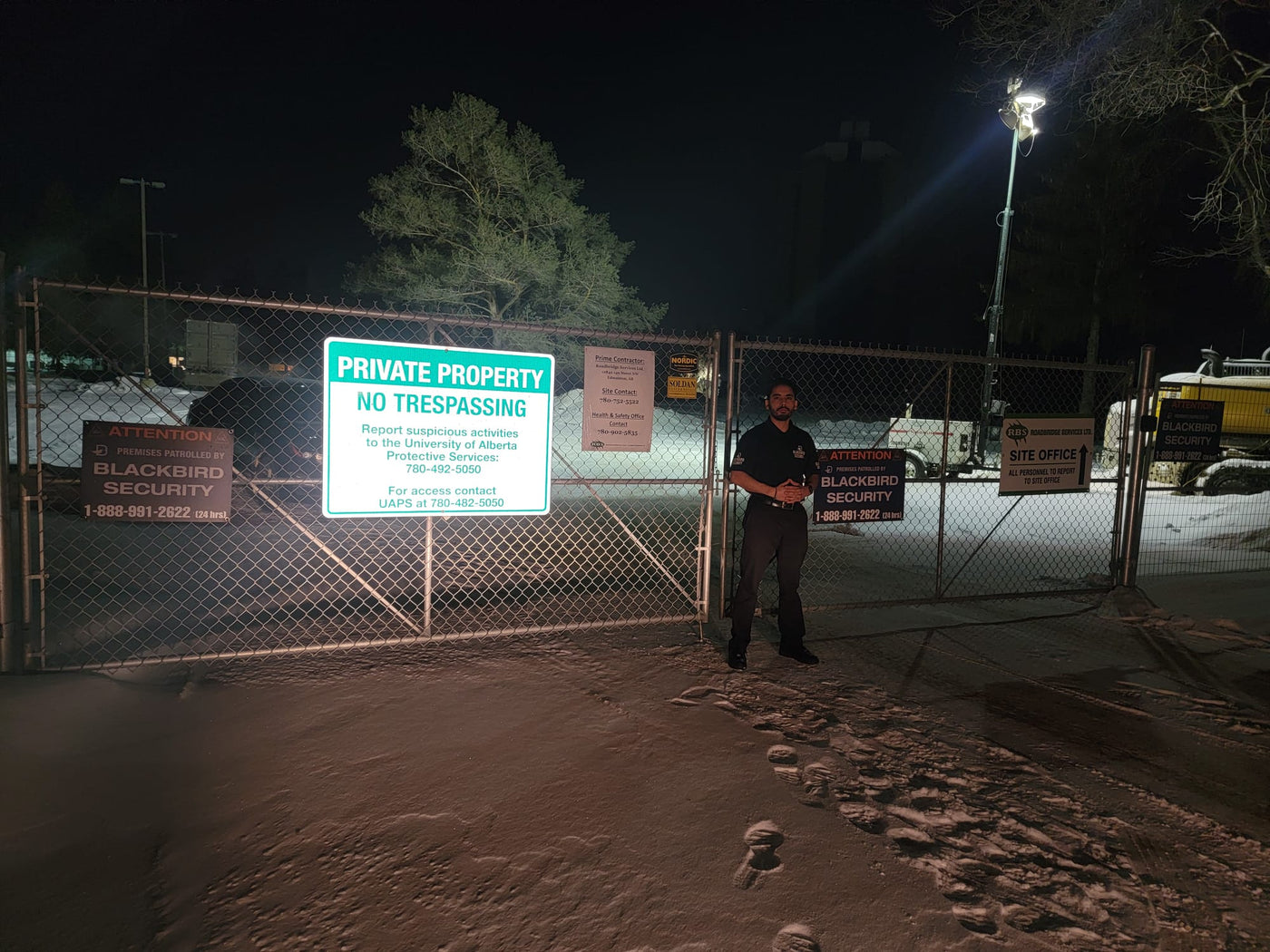 A uniformed security guard stands in front of a chainlink fence that showcases a sign reading "private property" during the nighttime hours.