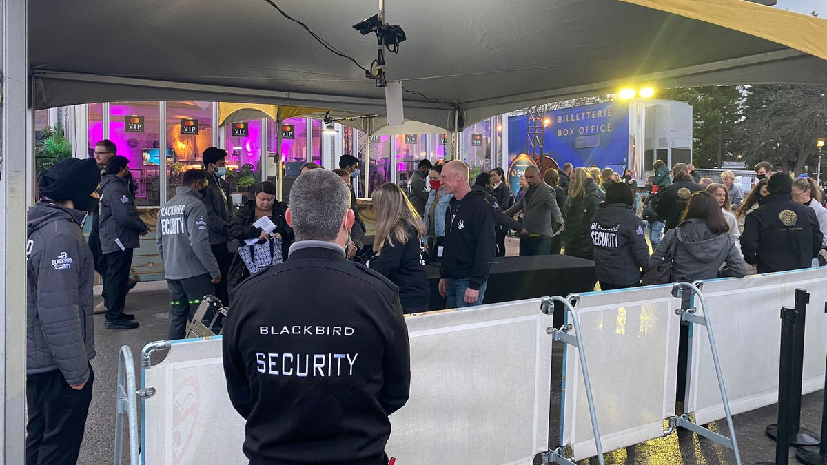 A number of Blackbird Security personnel are placed at various stations under a tent where a large crowd of people are entering an event. Some security guards are checking bags, others are keeping an eye on the crowd.