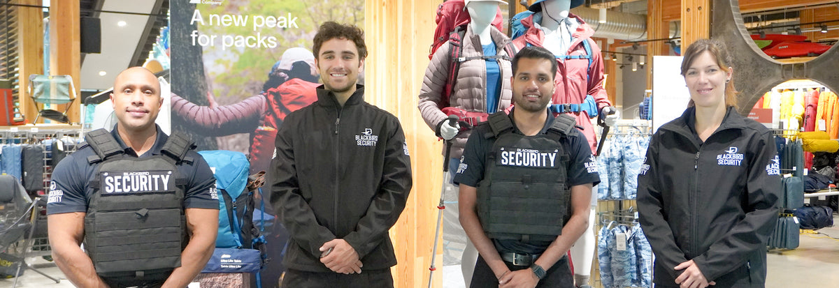 Four security guards are standing in front of a retail display of hiking gear. Two of the security guards are wearing black vests, two are wearing black jackets; all with Blackbird Security logos on them. They are all smiling and have a relaxed posture.