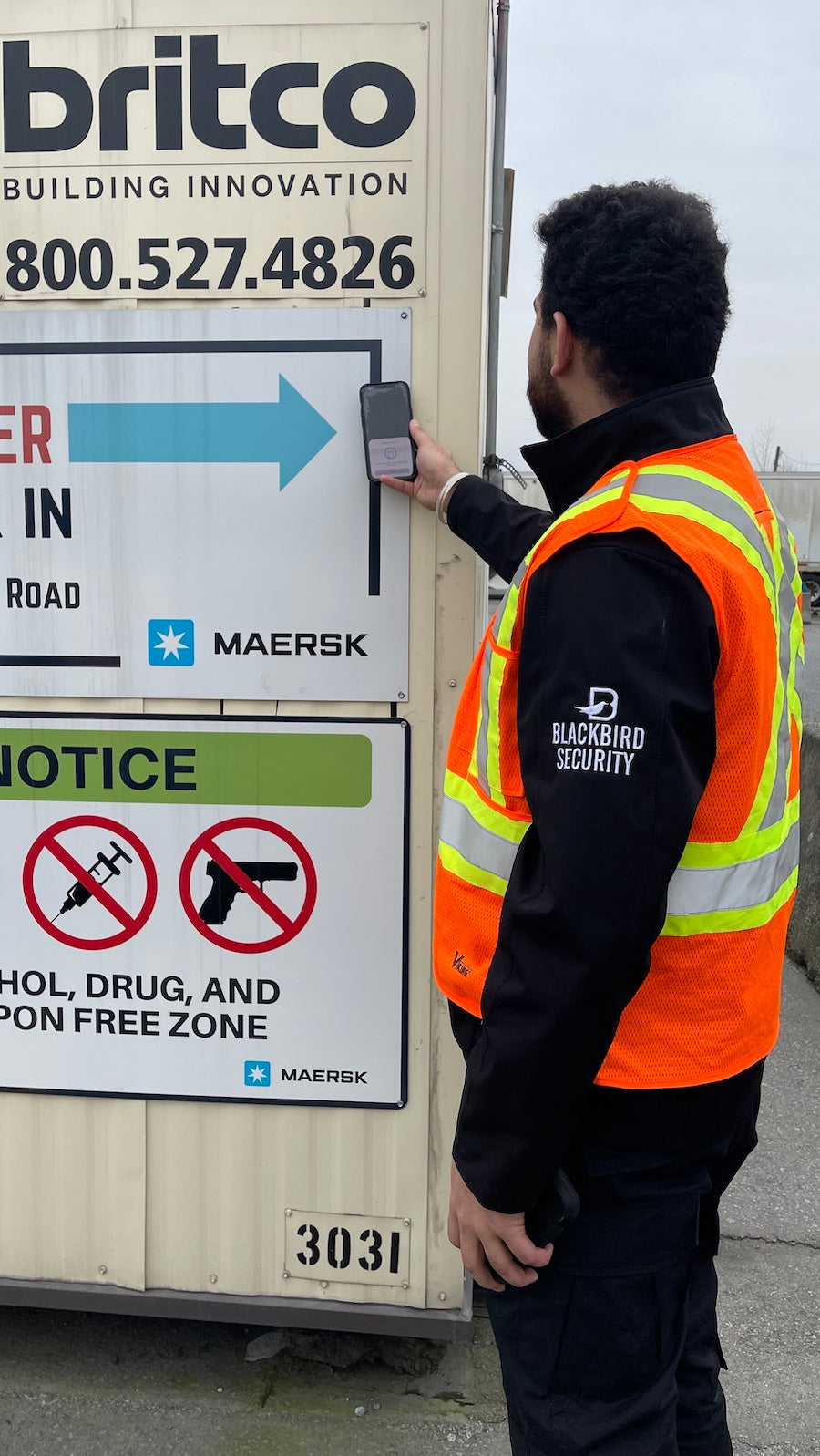 Uniformed security guard wearing a reflective vest is checking a shipping container at a construction site. They are holding a device up to the side of the container.