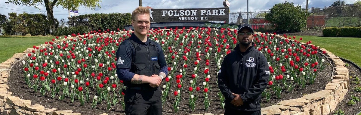 Two Blackbird Security personnel are standing in front of a large tulip garden with a sign that reads Polson Park, Vernon, BC at the top.