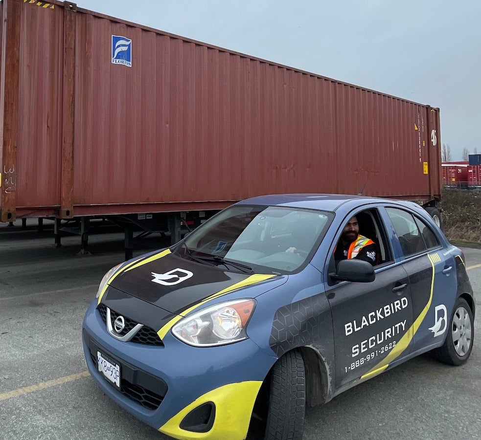 Security guard in a reflective vest is sitting inside the Blackbird Security mobile patrol car. It is in front of a large trailer in a parking lot.