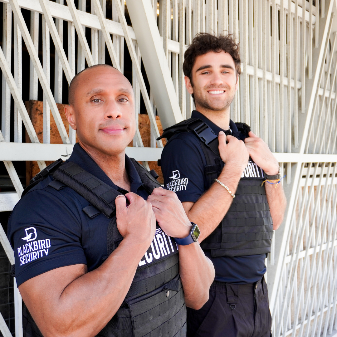 A pair of Blackbird security guard stand holding onto their vests stand outside of a metal gate