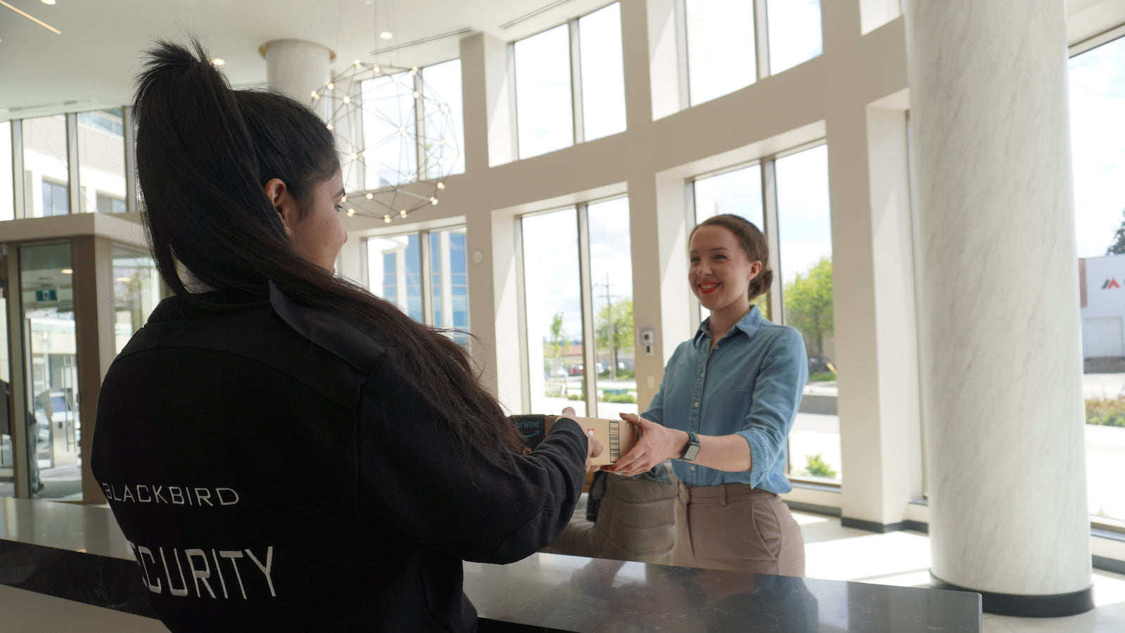 A Blackbird Concierge Security person with long hair is handing a package to a person on the other side of the counter. They are in a large foyer with a large pillar and windows behind them.