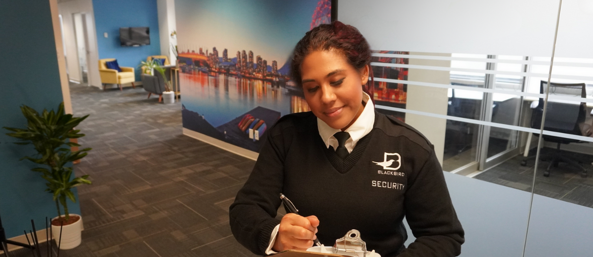 Blackbird Security guard is standing in an office looking down at a clipboard. They are writing on some paper with a pen