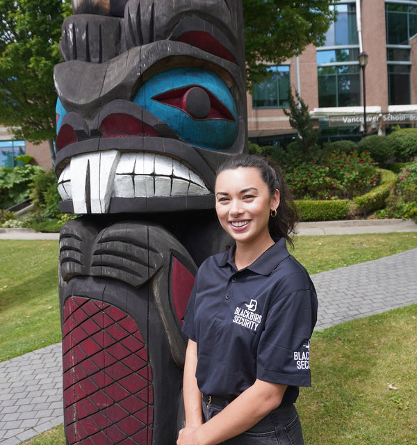 Blackbird security guard is standing beside a totem pole outside of the Vancouver School Board building. She is smiling, and it is a bright, sunny day.
