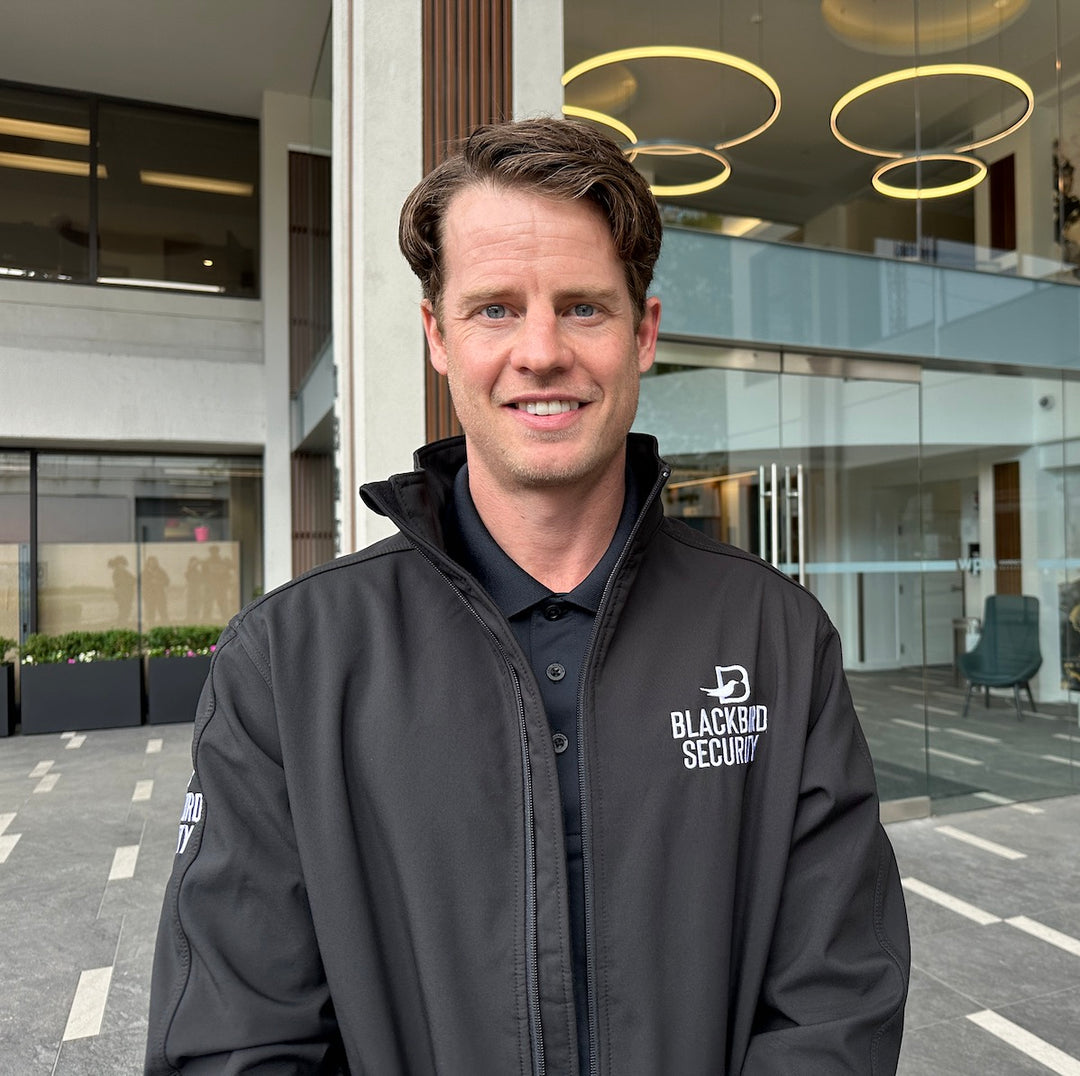 Blackbird Security guard wearing a black jacket with Blackbird logo stands in front of a conference centre entrance. They are looking into the camera and smiling.