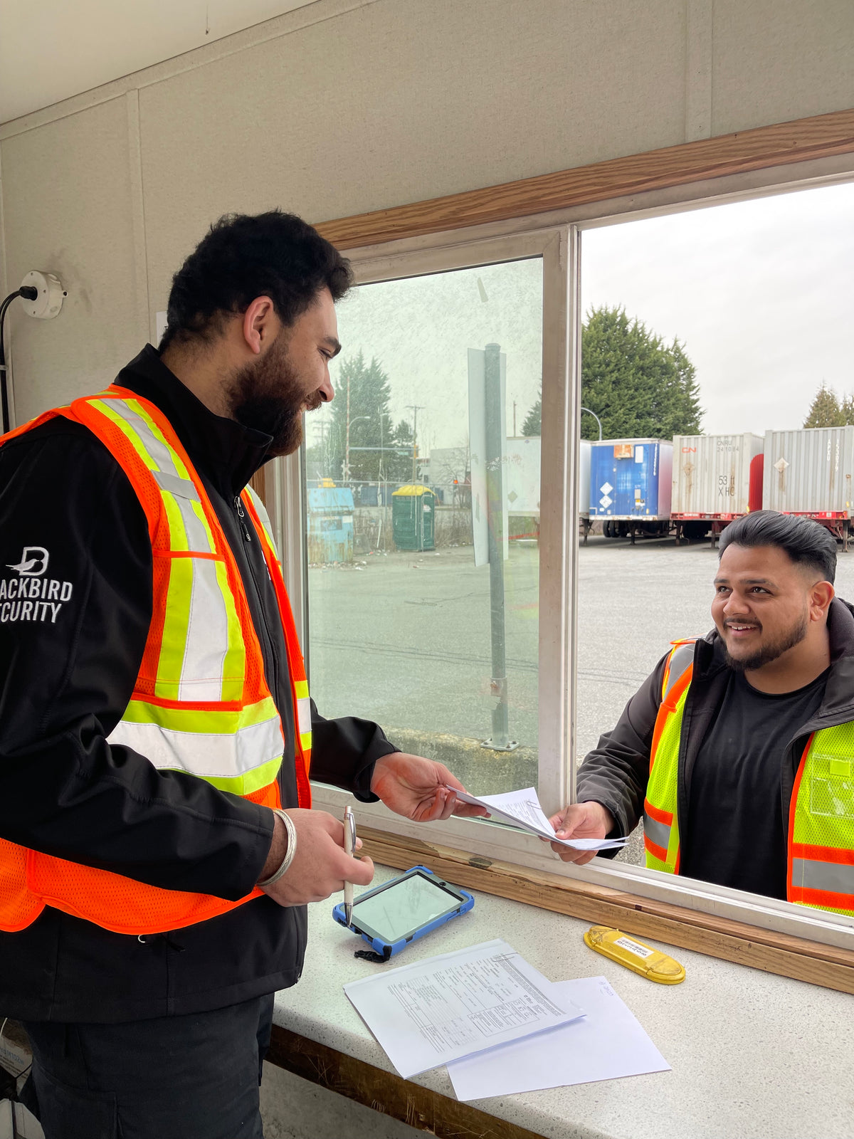 Blackbird industrial security guard stands at the window of a trailer speaking to someone outside. They are both wearing reflective vests.