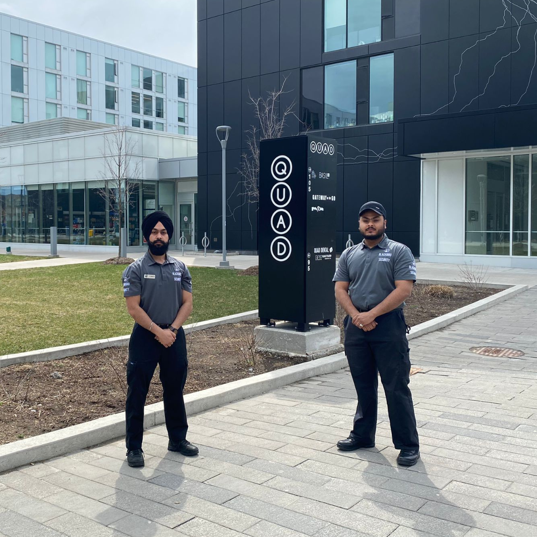 A pair of security guards standing on a school campus in front of a Quad sign