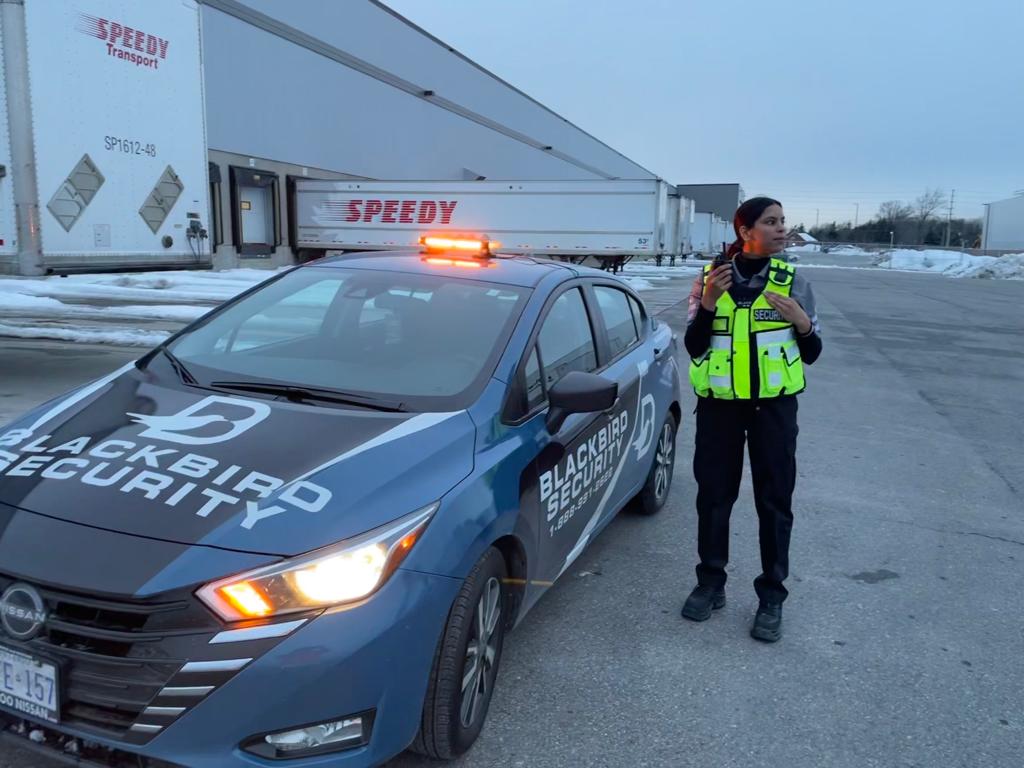 uniformed Blackbird Security guard wearing bright safety vest beside branded patrol car with lights on