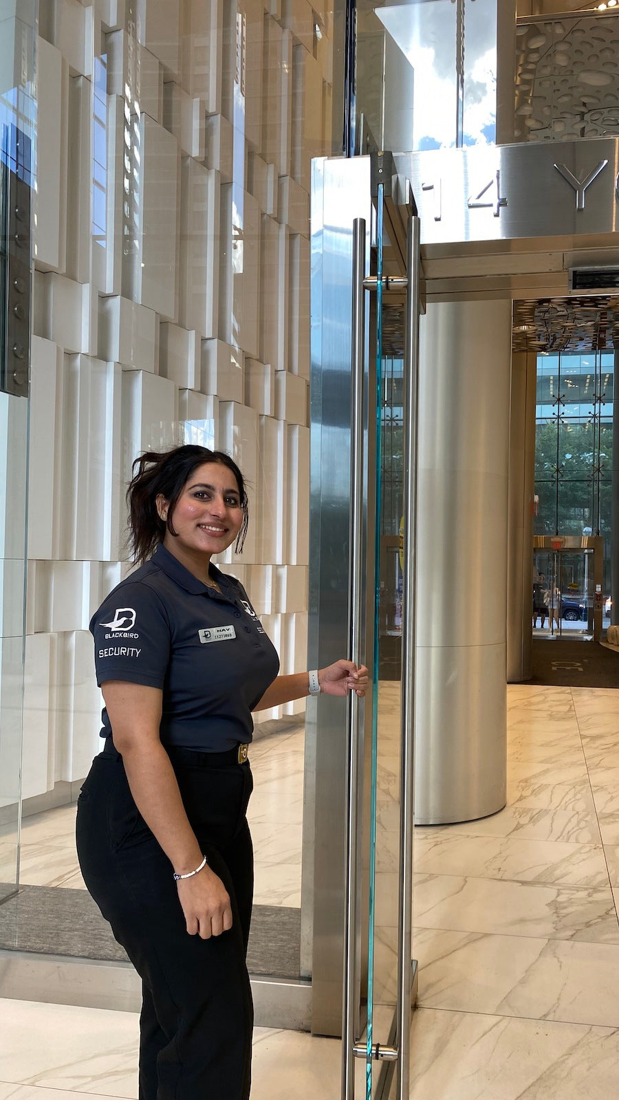 A female Blackbird Security guard holds open a large glass door at the entrance to a hotel building. She is smiling and appears friendly.