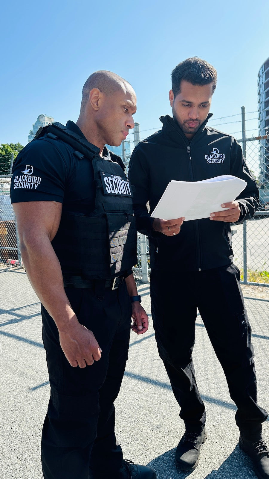 Two Blackbird Security guards are standing in front of a chain link fence looking at a paper document. It is a sunny day.