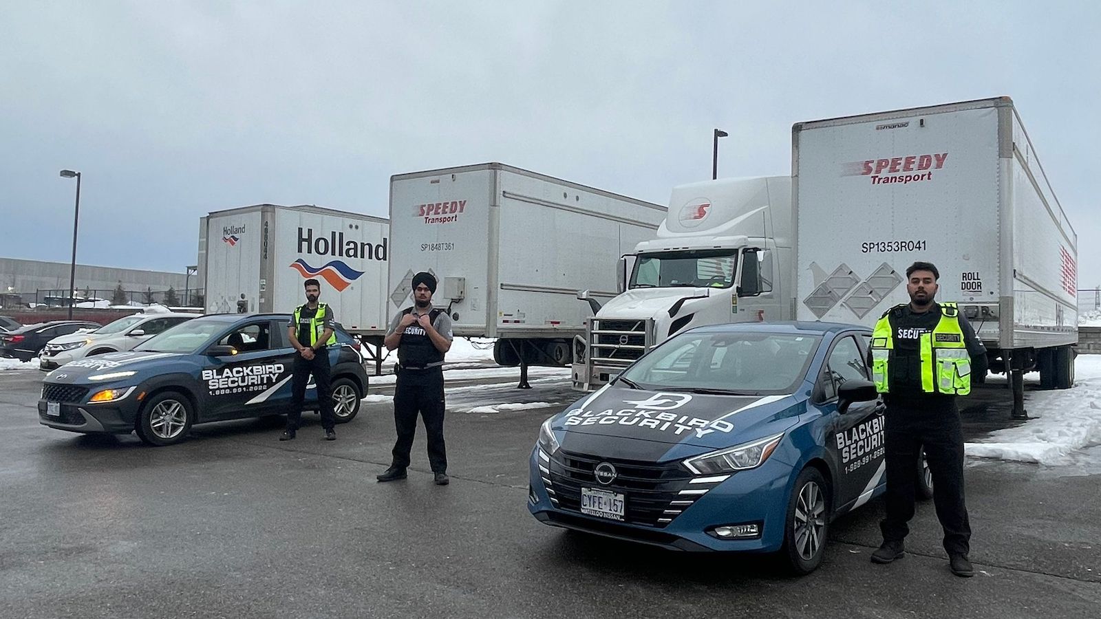 Blackbird Security guards are standing with their mobile patrol vehicles. They are in a parking lot with transport trucks parked behind them.